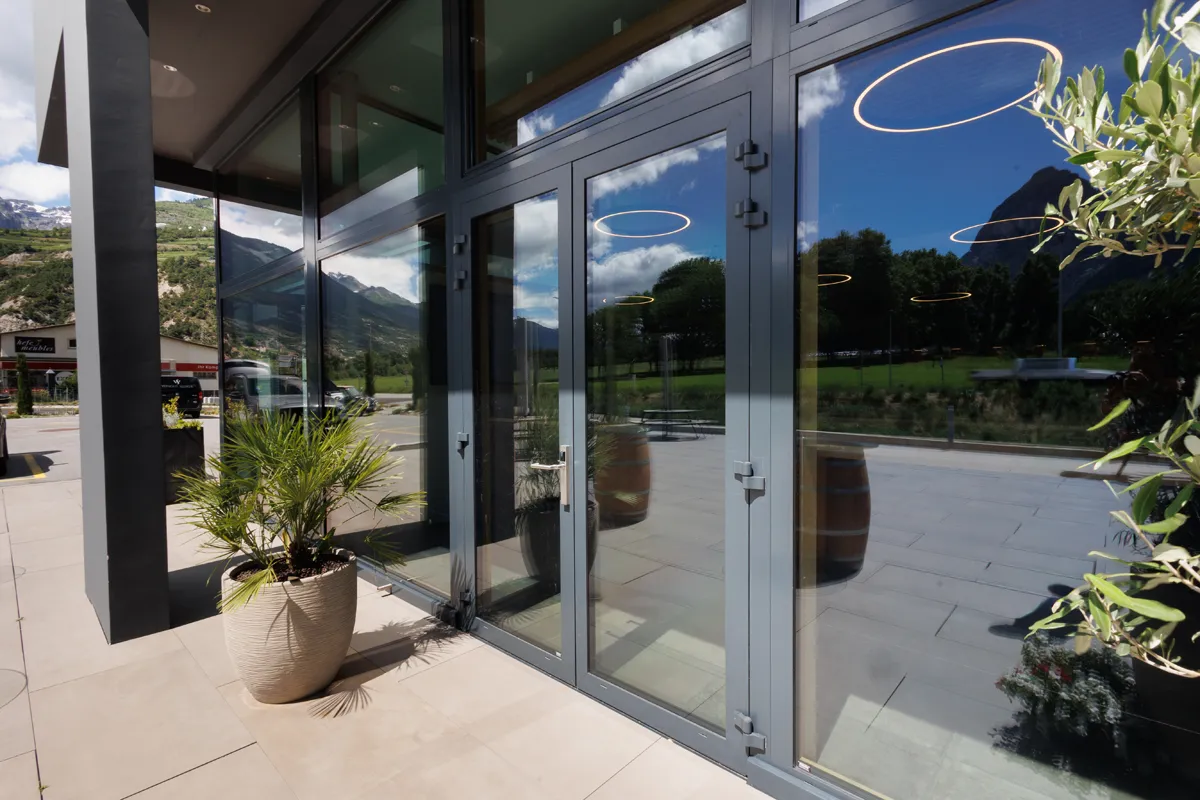Große Glasfassade mit Türen und Pflanzen vor moderner Gebäudefront im Oberwallis.